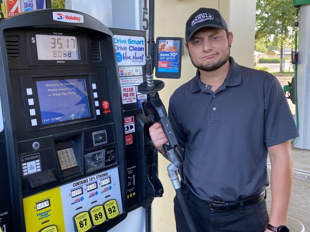 A man with a baseball cap on standing at a gas pump holding the gas nozzle.