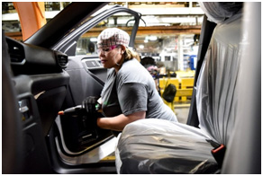 A woman wearing a gray tee short and a cap is holding a drill working on the inside of a Ford vehicle at a Ford plant.